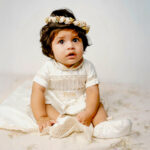 Baby in a vintage-style off-white baptism outfit with lace accents and the name embroidered, wearing a floral crown.