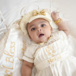 Baby boy in premium off-white baptism romper with gold embroidery, lying in decorated cradle.