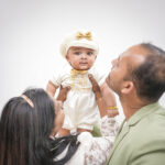 Baby lifted by parents, wearing premium personalized baptism romper and golden accessories.