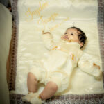 Baby boy in cream baptism dress lying on embroidered quilt with “Jayden Joseph
