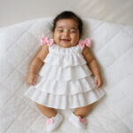 Smiling baby girl wearing Alice Sp white baptism dress with pink bows, lying on a quilted bed, showcasing premium satin and hacoba fabric.