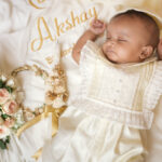 A peacefully sleeping baby dressed in a premium off-white christening romper with lace detailing, lying on a personalized baptism quilt embroidered with the name “Akshay,” accompanied by a floral crown and decorated baptism candle.