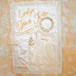 Personalised baby baptism dress, bonnet, and floral crown displayed on embroidered quilt