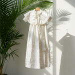 An exquisite white satin heirloom baptism gown for a baby girl, featuring a detachable capelet with delicate lace trim and cross embroidery, hanging on a wooden hanger in soft natural light.