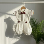 An elegant off-white satin baptism romper for a baby boy, featuring a rich burgundy bowtie, lace bib detailing, and matching burgundy piping on the capped sleeves, hanging on a wooden hanger.