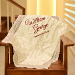 A luxurious personalized ivory satin baptism blanket featuring a wide lace border, custom embroidered with the name 'William George' in elegant burgundy thread, draped over a wooden rocking chair.