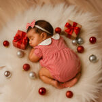 Baby sleeping in a red striped romper with white collar surrounded by Christmas ornaments