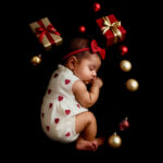 Sleeping baby in a white romper with red heart prints surrounded by Christmas ornaments