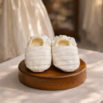 Off white handcrafted satin baby baptism booties displayed on a wooden stand